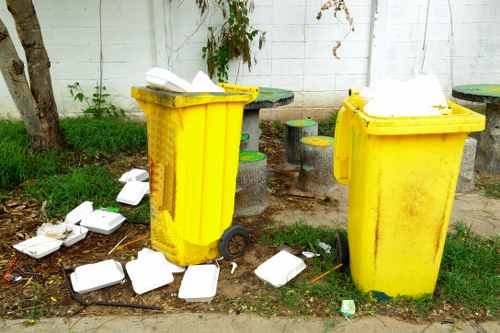 Two large yellow wheelie bins with black wheels are positioned outdoors on a pavement, placed side by side in front of a white wooden fence. The bin on the left has a partially open lid with white polystyrene food containers and packaging scattered on the ground around its base. The bin on the right is closed, with some white foam packaging visible on top of its lid. The area behind the bins features a small garden space with grass, a tree trunk, overgrown plants, and a few concrete tables and stools with yellow and green painted surfaces. The scene appears to be part of a residential or commercial waste collection area, with natural daylight illuminating the scene, emphasizing the bright yellow of the bins and the scattered waste materials. This visual is representative of rubbish collection points that commercial waste removal services, such as Commercial Waste Temple, might handle in the local area around Temple.