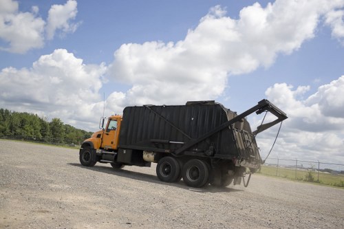 A large industrial waste collection truck parked on a gravel surface, with a black, metal waste container mounted on its chassis. The container is partially tipped at an angle, indicating unloading or loading activity, with its hinged rear door slightly open and a metal lever visible. The truck's cab is orange, contrasting with the dark container, and the scene is set outdoors under a partly cloudy sky with white clouds and patches of blue. The background shows open fields and some distant trees, suggesting a rural or semi-rural environment, typical of commercial waste collection services near Temple, with no other vehicles or objects in immediate view. The truck and container are clean and well-maintained, relevant to professional rubbish removal by Commercial Waste Temple in the local area.