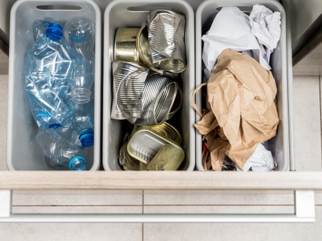 The image shows three separate grey plastic waste bins filled with different types of rubbish, situated on a tiled floor, likely in a commercial or residential setting. The left bin contains empty, crushed plastic water bottles with blue caps and labels, arranged loosely and spilling slightly over the sides. The middle bin holds a pile of crushed aluminum cans with a shiny metallic finish, many stacked unevenly, some leaning against the sides. The right bin contains crumpled paper and brown kraft paper bags, with some wrapping and packaging material loosely layered. The bins are positioned on a light-colored tiled surface with a neutral background, emphasizing the different materials ready for rubbish removal. The scene illustrates typical waste segregation for professional rubbish collection services and highlights the importance of proper disposal for recycling and waste management, relevant to commercial waste removal in Temple. Commercial Waste Temple provides these services to help maintain clean and clutter-free premises in the local area.