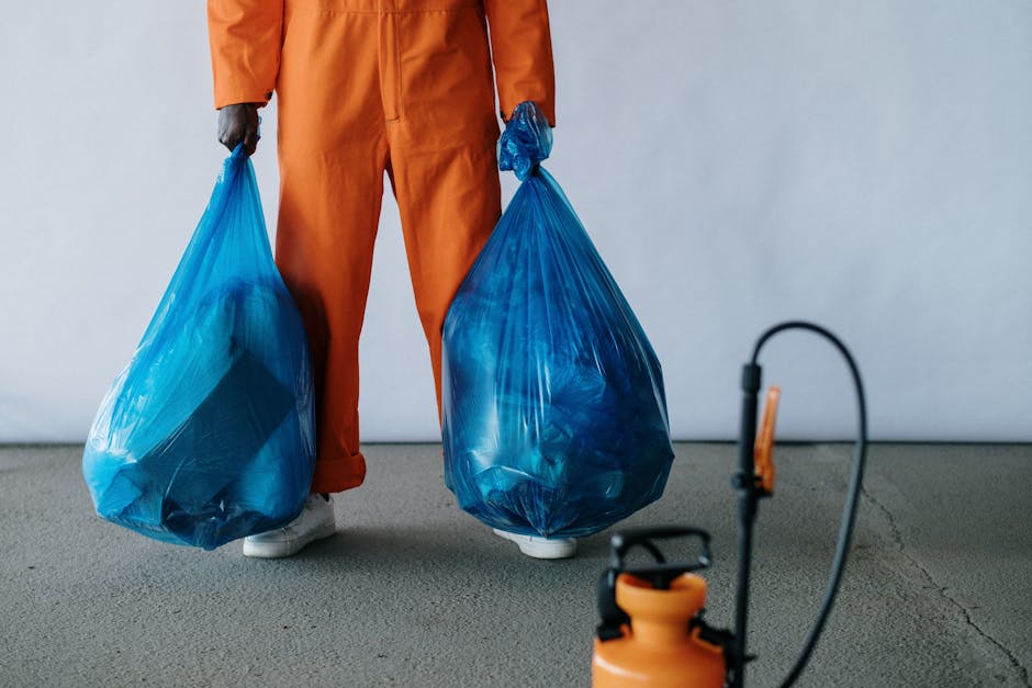 A person dressed in an orange work uniform is standing on a concrete floor, holding two large blue plastic rubbish bags, one in each hand. The bags are semi-transparent, revealing waste materials insi